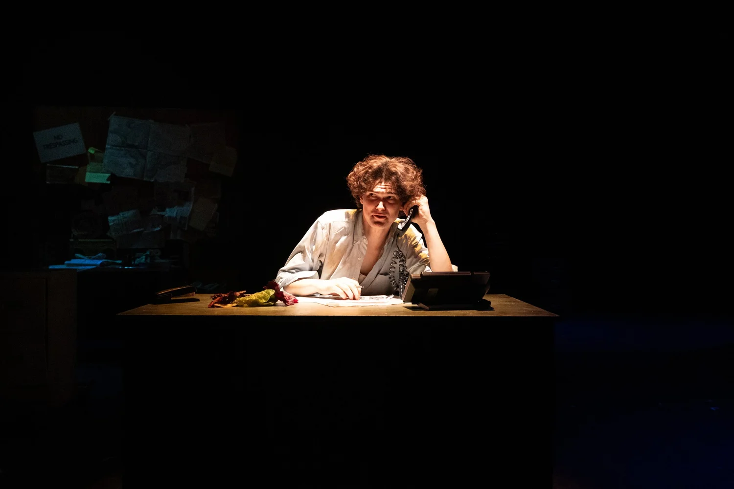 Photo of an actor sitting at a desk and holding a telephone.