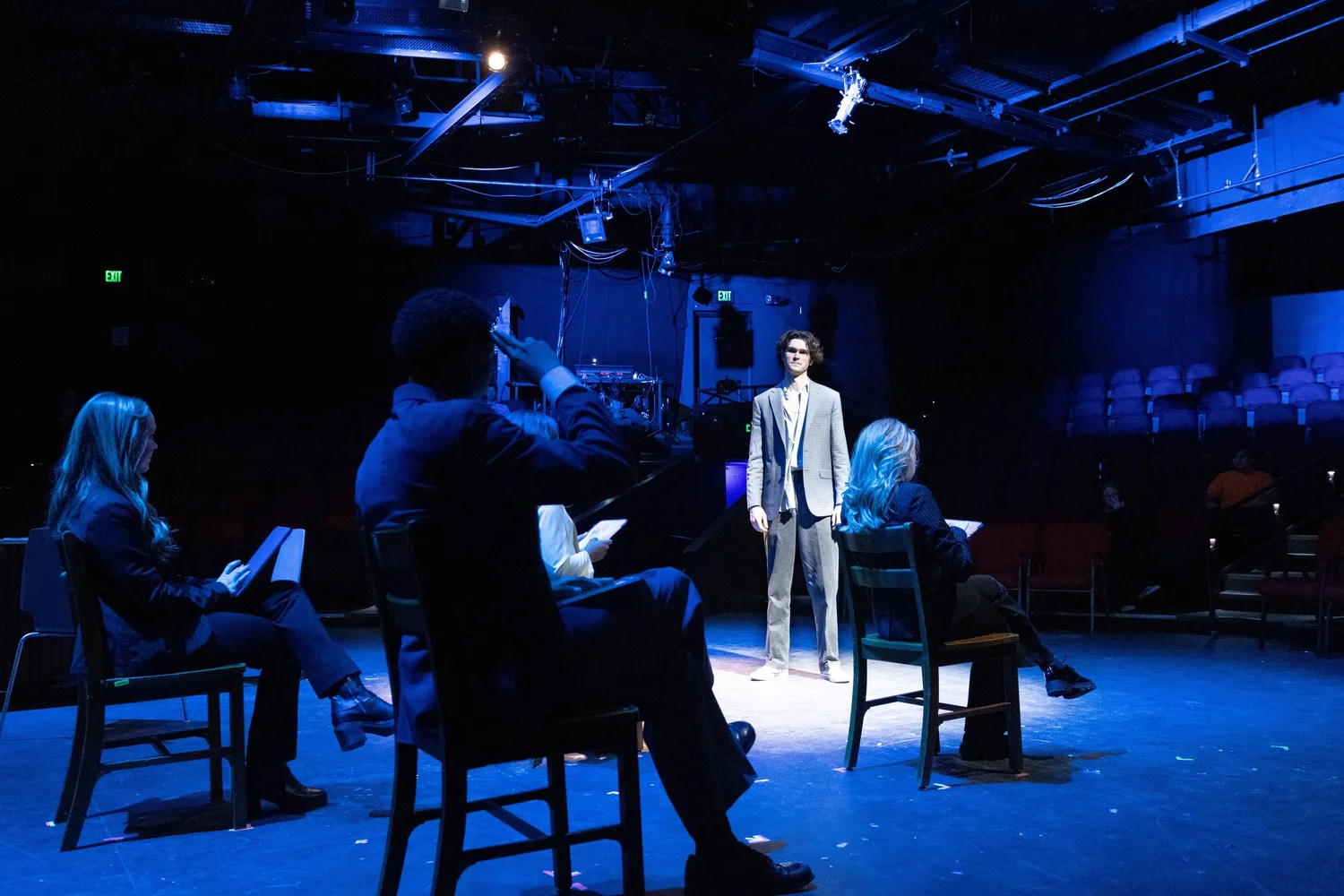 Photo of actors seated in the foreground with an actor standing under the spotlight in the background.