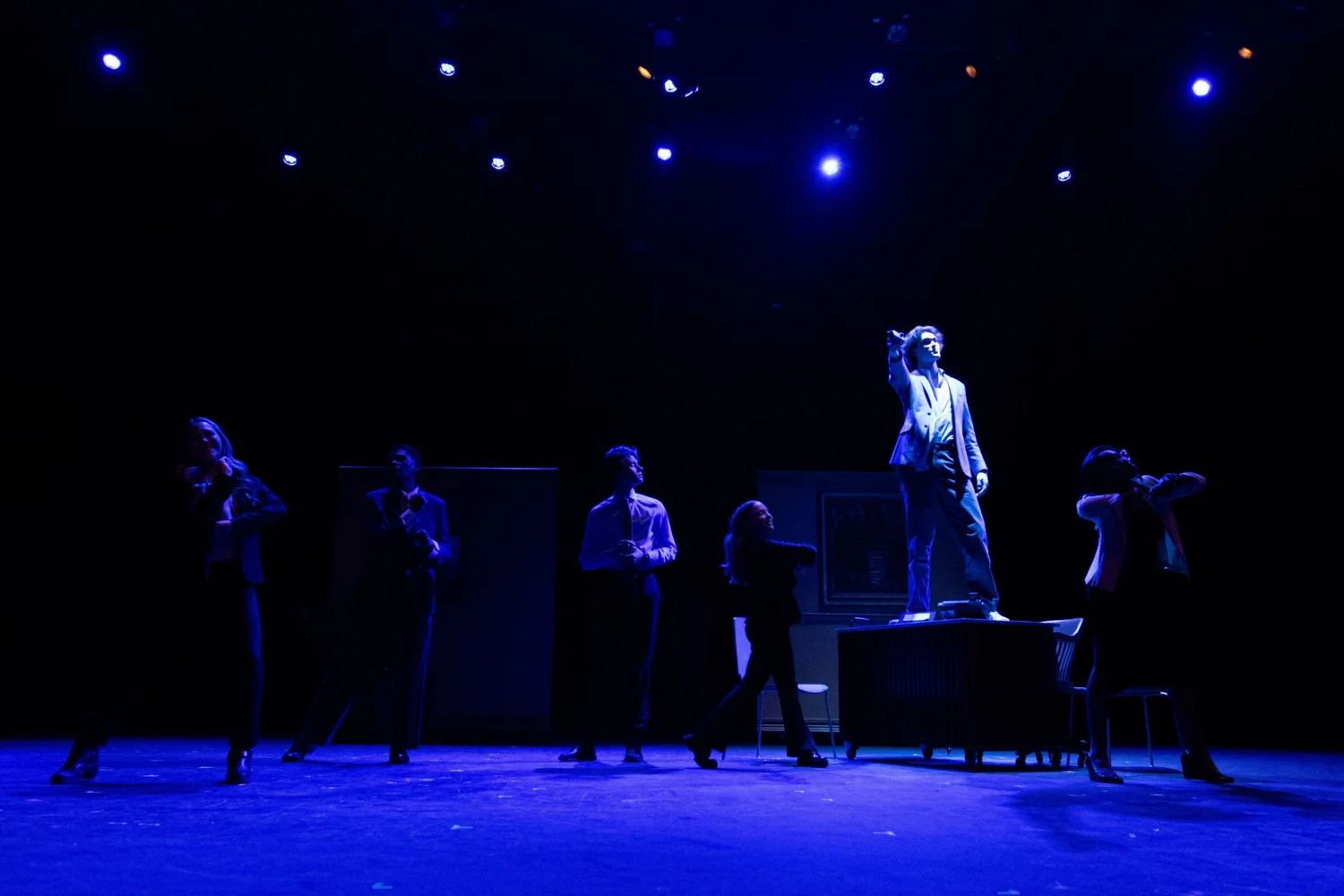 Photo of an actor standing on top of a desk while other actors on the ground dance.